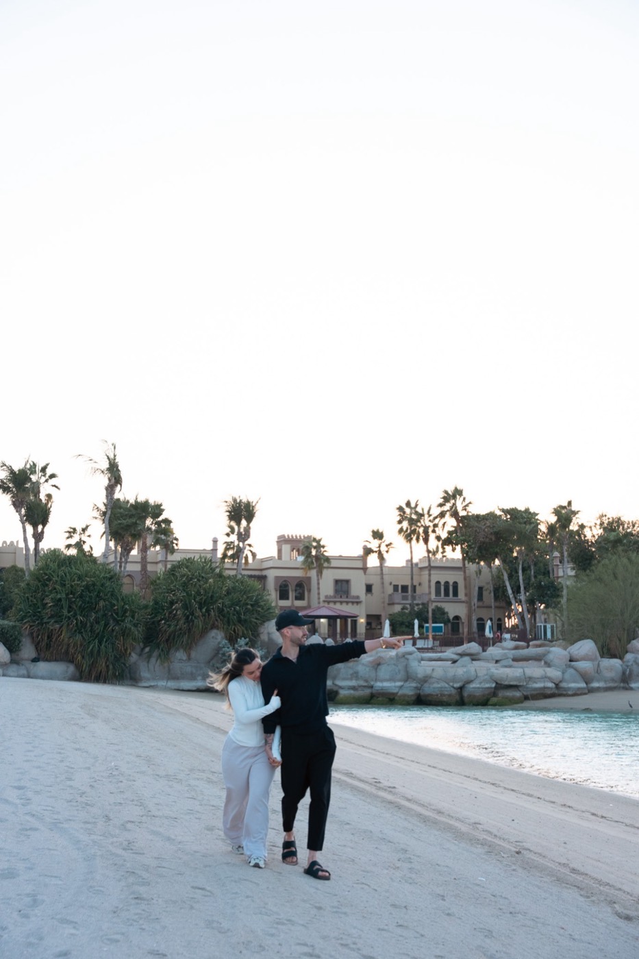 Valentina y Javi caminando abrazados por la playa, él señalando hacia delante